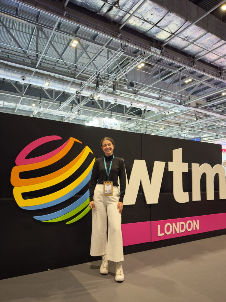 A woman wearing a black top and white pants stands smiling in front of a large "WTM London" sign at an indoor convention or event space.