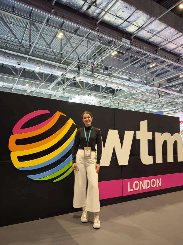A woman wearing a black top, white pants, and white boots stands smiling in front of a WTM London event sign inside a large convention center during November in London.