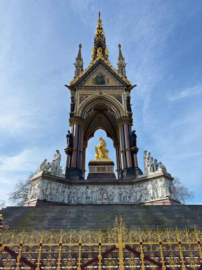 The image shows the ornate Albert Memorial in London, with a golden statue of Prince Albert seated under a decorated Gothic canopy, surrounded by white marble figures and intricate iron fencing in the foreground.