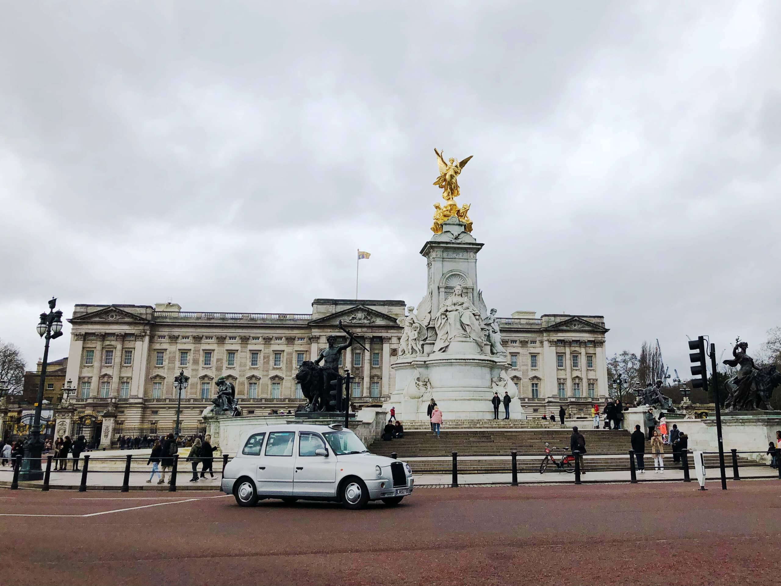 Buckingham Palace under a cloudy sky with the Victoria Memorial in front, a white taxi driving by, and people walking around the monument and plaza. Visiting Buckingham Palace is one of the most iconic things to do in Westminster London