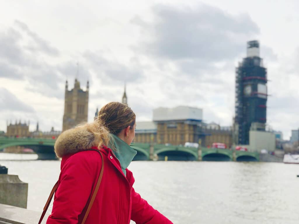 A person in a red coat stands by the River Thames, looking towards the Palace of Westminster and Big Ben, which is covered in scaffolding. The sky is cloudy and the scene is in London, England. Crossing the Westminster Bridge is one of the best things to do in Westminster London