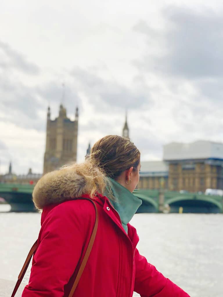 A person in a red coat stands by the River Thames, looking towards the Palace of Westminster and Big Ben, which is covered in scaffolding. The sky is cloudy and the scene is in London, England.
