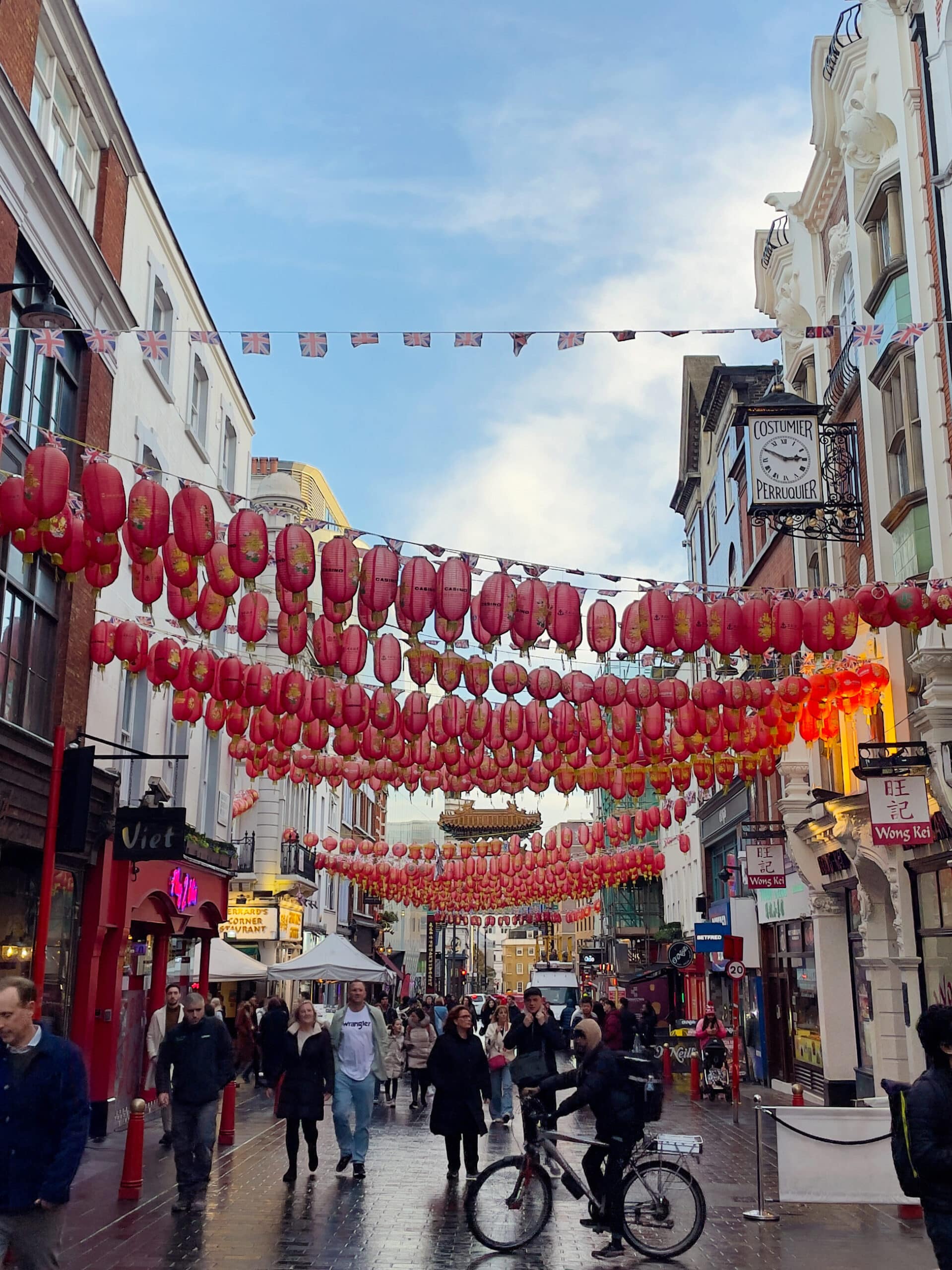 A busy street scene in London's Chinatown with red lanterns hanging overhead, people walking and cycling, and shops with colorful signs lining the wet pavement under a partly cloudy sky.