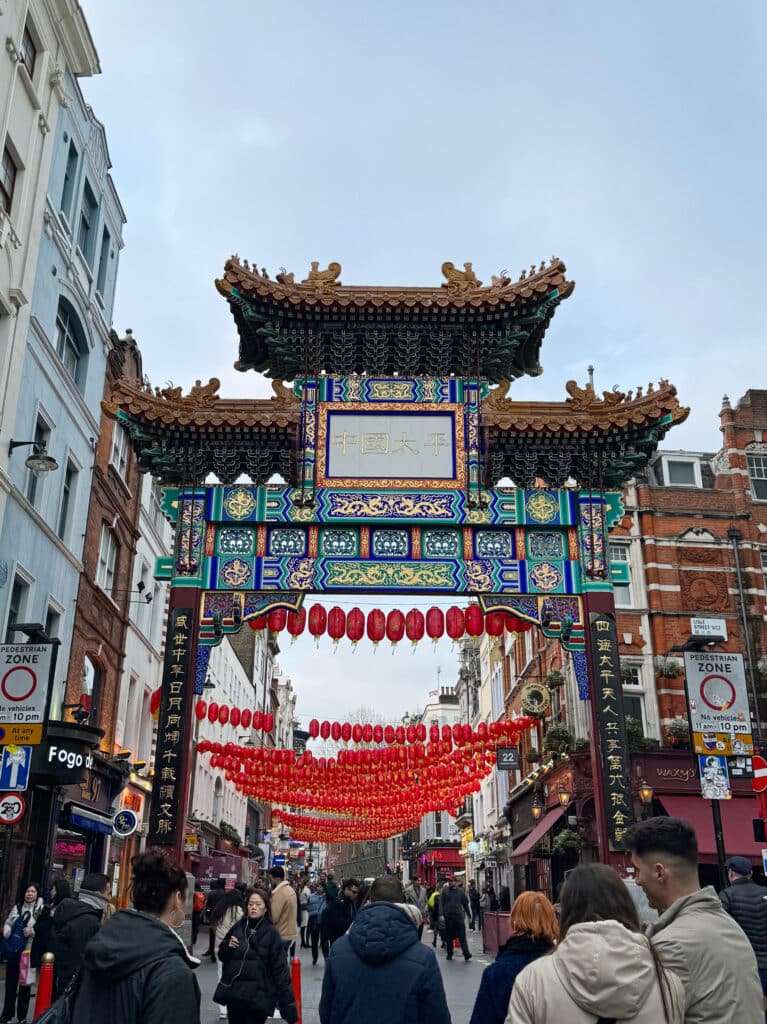 Chinatown's colorful Chinese gate with ornate patterns and gold details stands over a busy street decorated with red lanterns. People walk below, surrounded by shops and tall buildings on a cloudy day.