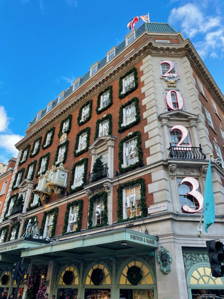 A historic brick building decorated with festive greenery and large numbers "2023" on its facade, under a blue sky. Experience London in November at Fortnum & Mason, where its iconic clock and arched windows welcome visitors.