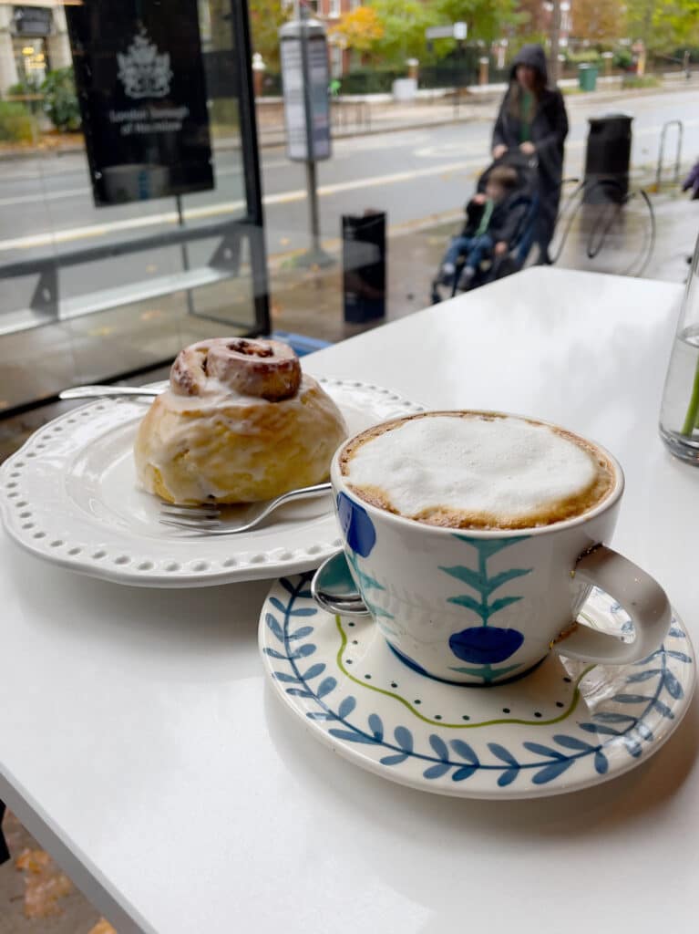 A cappuccino in a patterned cup and saucer sits beside a glazed cinnamon roll on a white plate, on a café table by a window in gluten free London, with a blurred street and person pushing a stroller outside.