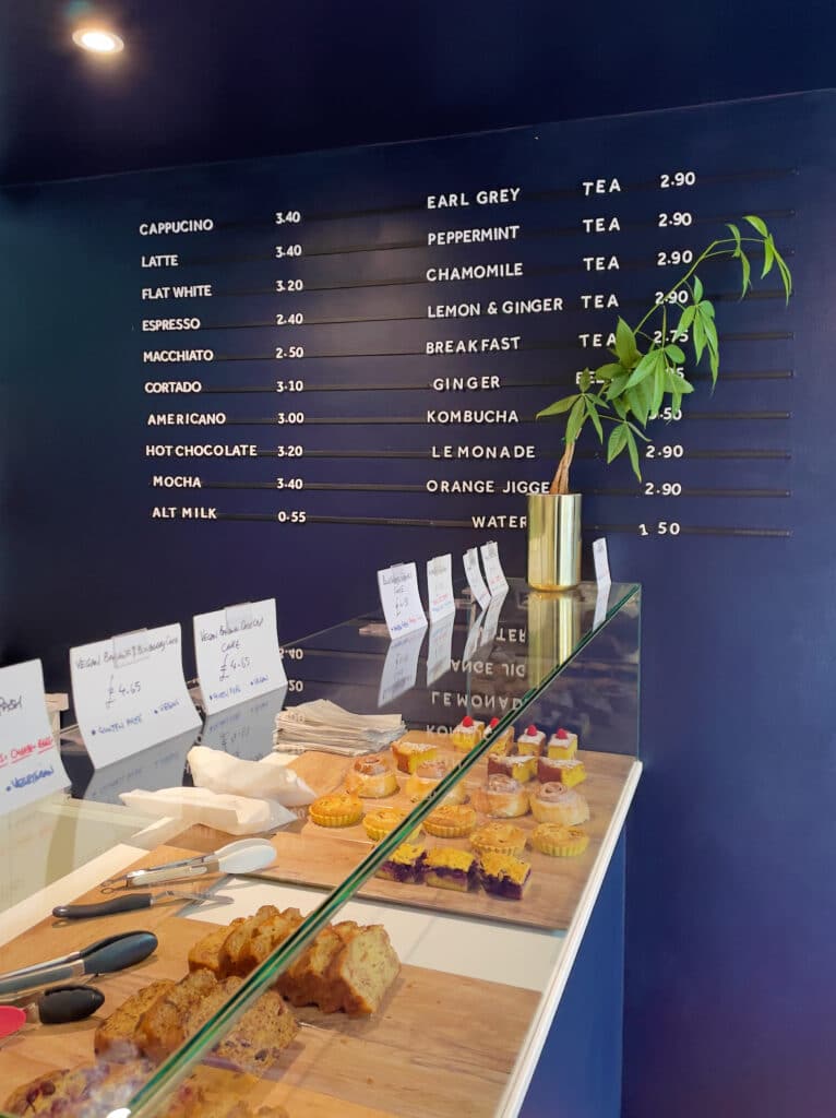 A cafe counter in London with gluten free pastries and cakes on display, set before a dark blue wall menu listing coffee and tea options with prices, and a small plant in a gold vase atop the counter.