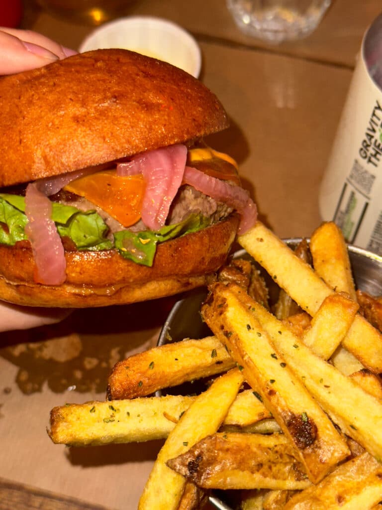 A hand holds a burger with lettuce, cheddar cheese, and pickled onions next to a bowl of seasoned French fries on a brown tabletop, perfect for those seeking gluten free options in London.