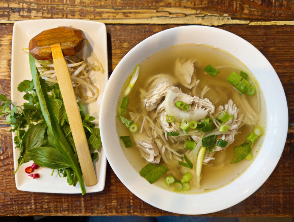 A bowl of Vietnamese pho with sliced chicken, green onions, and noodles in clear broth—ideal for those seeking gluten free London options—served with fresh herbs, bean sprouts, chili, and a wooden spoon on a rustic wooden table.