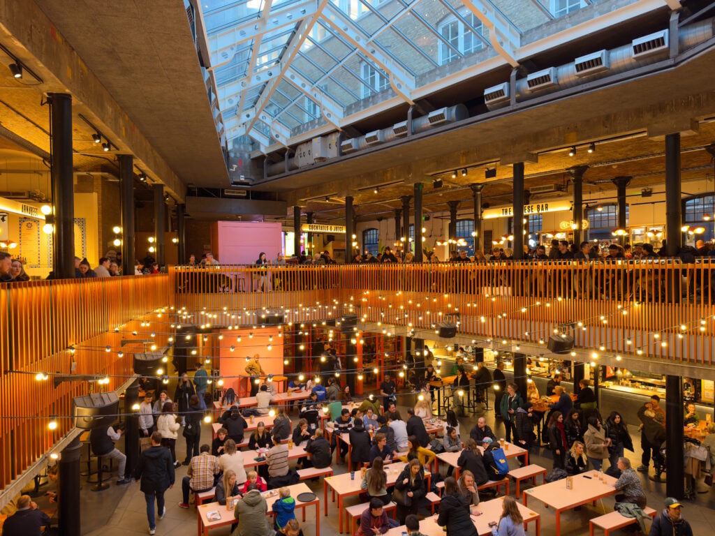 A busy indoor food market, Seven Dials Market, with people sitting at communal tables on the ground floor and more people gathered on the upper level. String lights hang from the ceiling, and a large skylight lets in natural light.