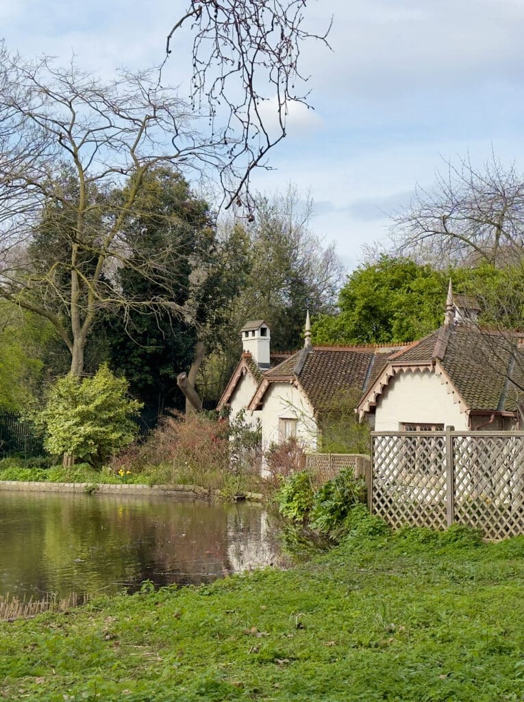 A small white cottage with a red-tiled roof sits beside a pond, surrounded by trees, bushes, and grass, under a partly cloudy sky. A wooden lattice fence runs along the right side of the image. Visiting the St. James's Park is one of the best things to do in Westminster London