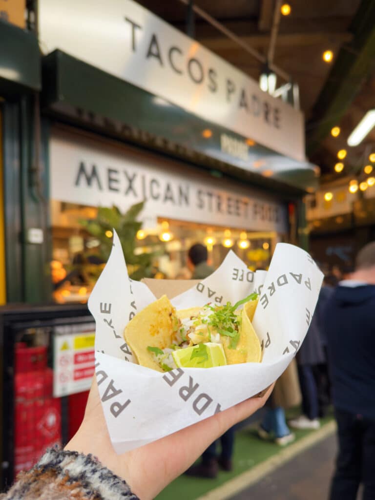 A hand holds a taco wrapped in branded paper in front of a food stall with signs reading "Tacos Padre" and "Mexican Street Food," popular for serving gluten free London street eats, with people standing nearby.