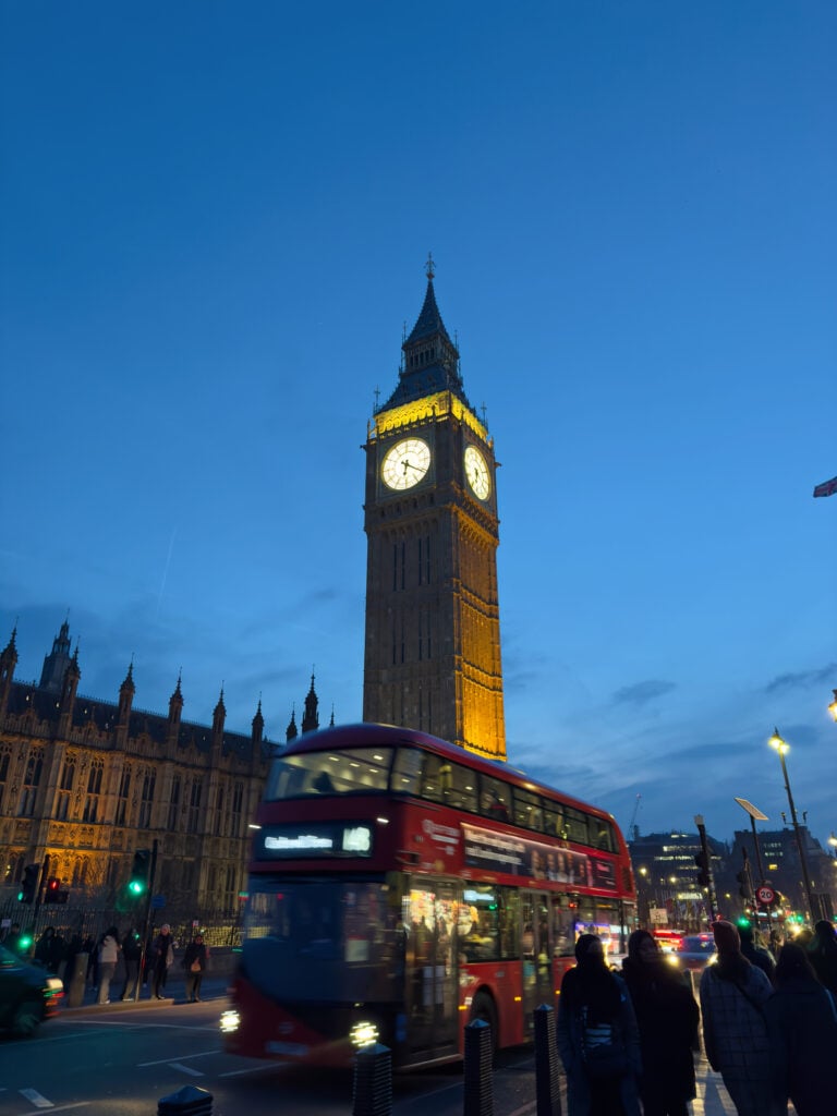 Big Ben clock tower illuminated at dusk in London, with a red double-decker bus passing by and people walking along the street in the foreground.