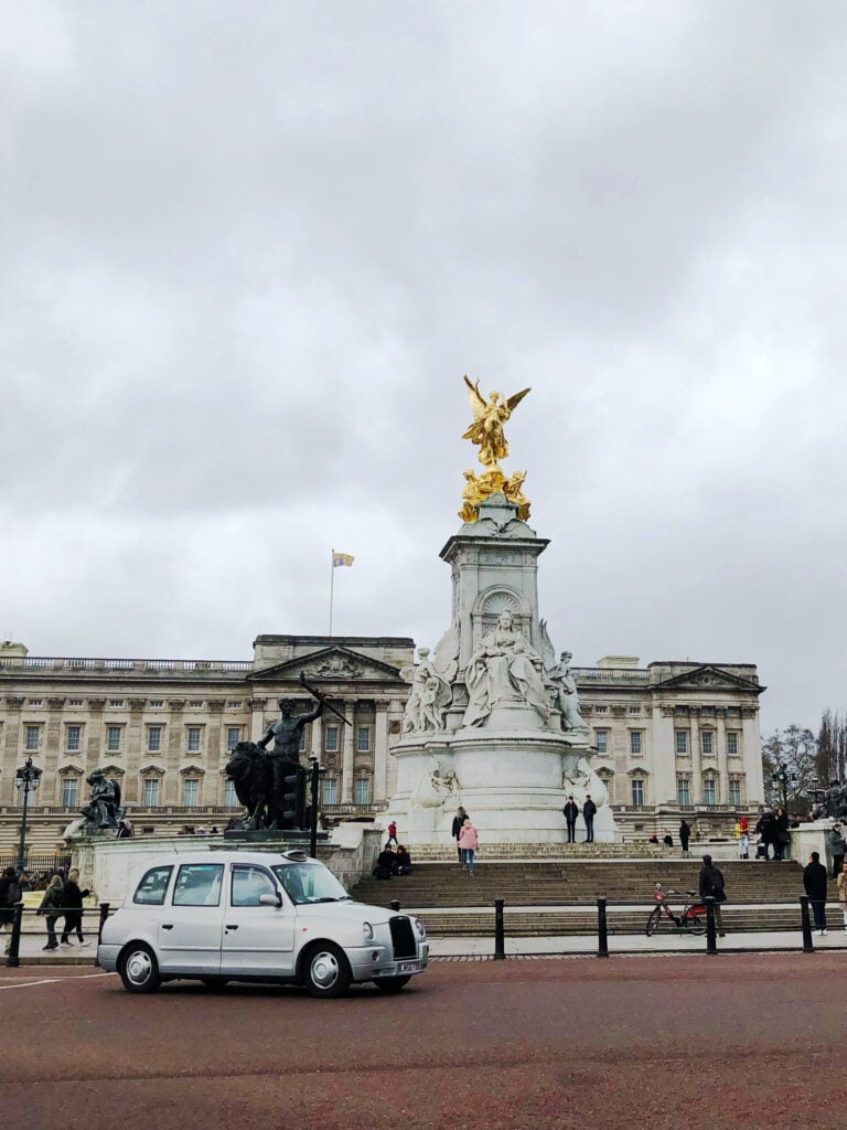 Buckingham Palace under a cloudy sky with the Victoria Memorial in front, a white taxi driving by, and people walking around the monument and plaza.