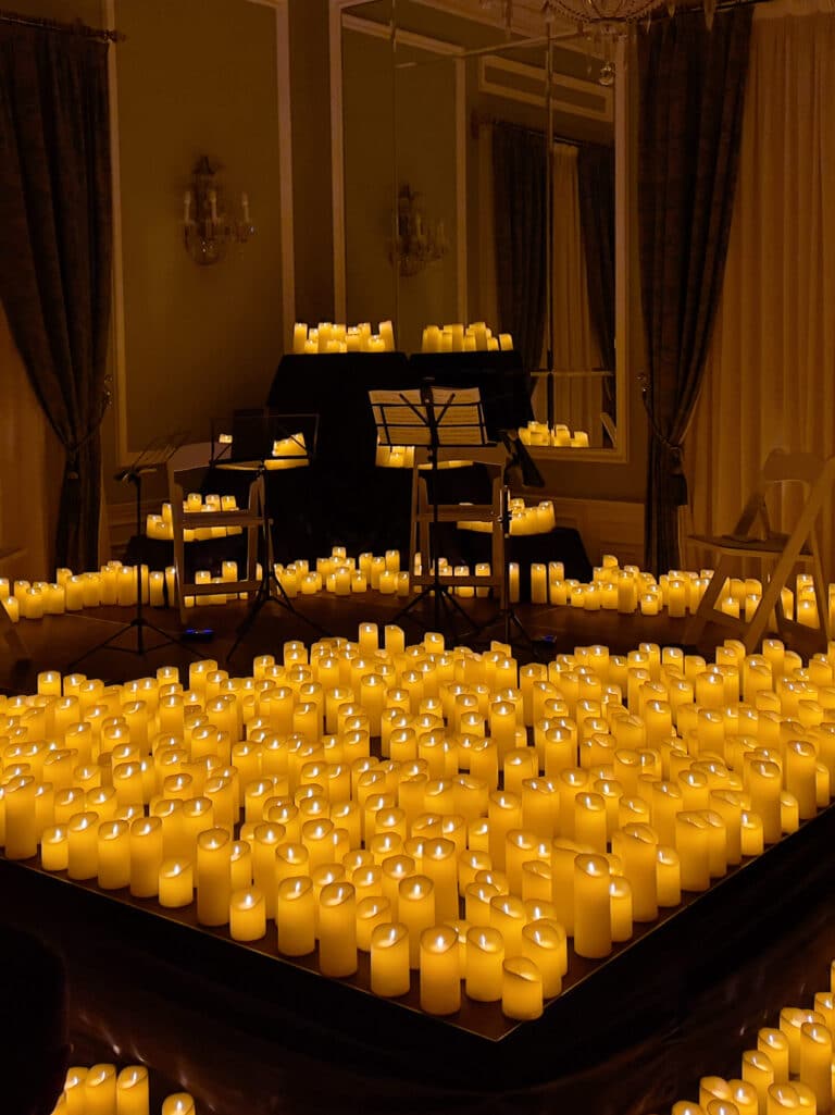 A stage surrounded by hundreds of glowing candles features empty music stands, a piano, and chairs, creating a warm, intimate atmosphere in a dimly lit, elegant room with curtains and mirrors.