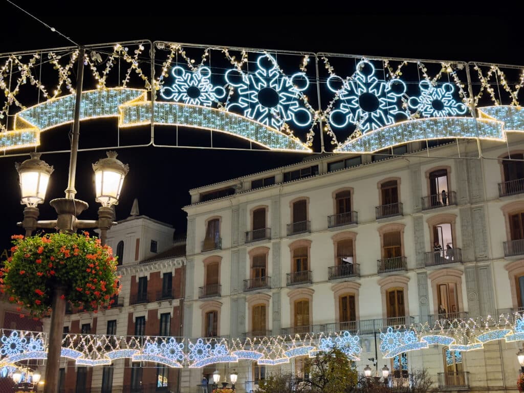 Festive holiday lights with blue snowflake designs hang above a city street at night in Granada in December, with illuminated buildings, classic street lamps, and a flower basket adding to the charming winter atmosphere.