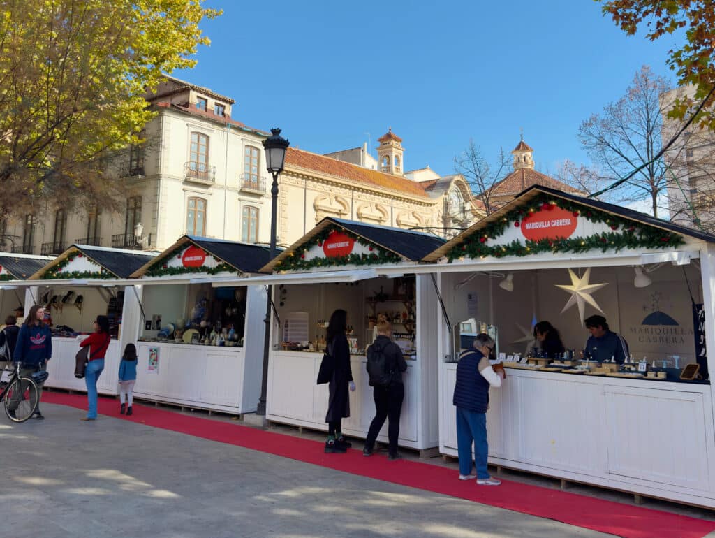 Outdoor holiday market in Granada in December with white wooden stalls decorated with garlands. People browse vendor booths along a red carpet, set against historic buildings and colorful autumn trees.