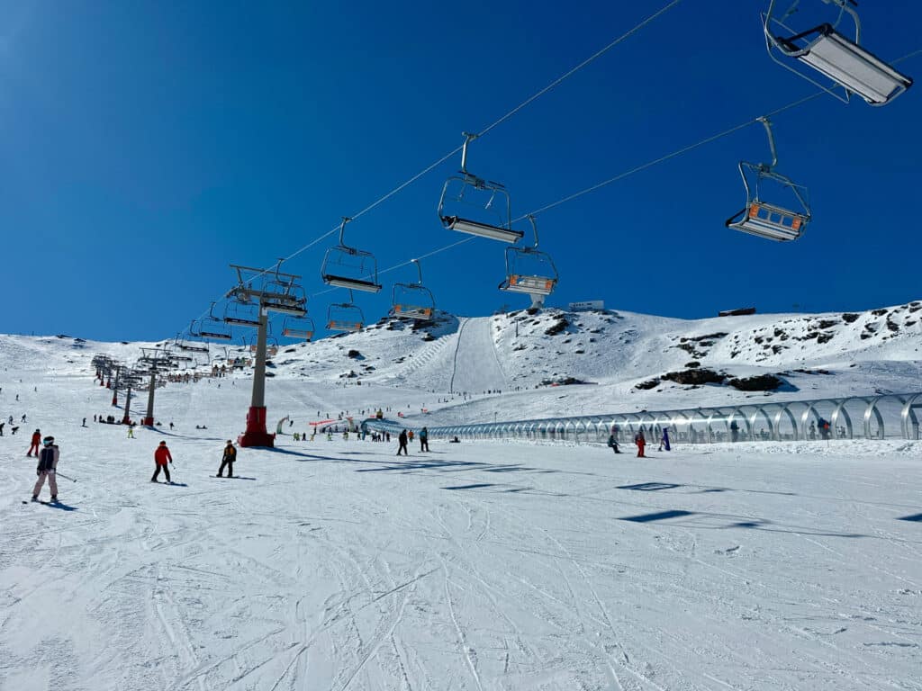 Skiers and snowboarders enjoy a sunny day on a snowy mountain in Granada in December as chairlifts move overhead. The clear blue sky contrasts with the white snow, with people visible near the ski lifts and a covered conveyor.