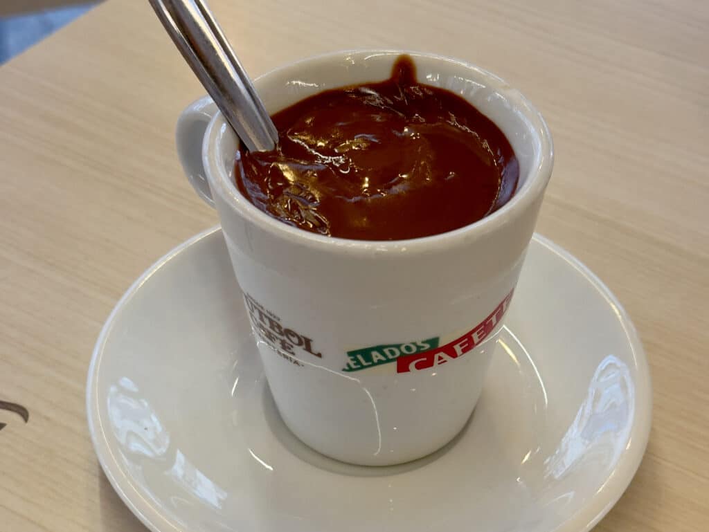 A white cup filled with thick hot chocolate, with a spoon inside, sits on a matching saucer on a light wooden table—just the kind of cozy treat you'll find in Granada in December. The cup has colorful labels with partial text visible.