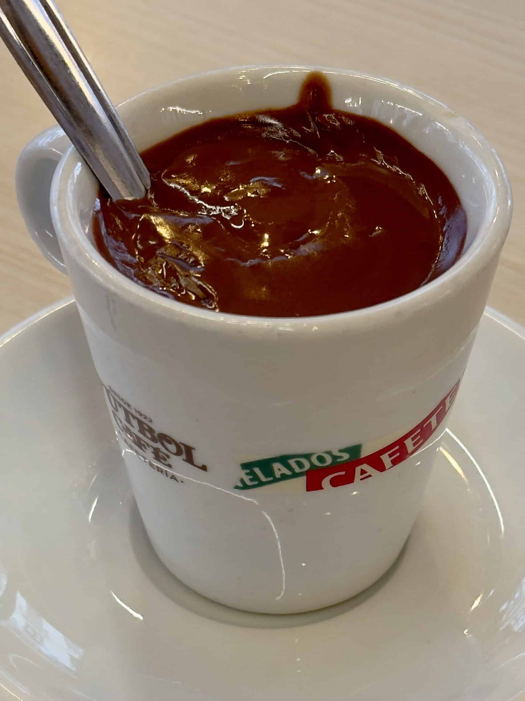 A white cup filled with thick hot chocolate sits on a matching saucer, with a metal spoon inside. The cup has colorful text on it and rests on a light wooden table.