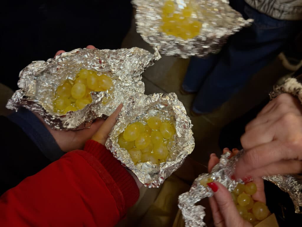 Several people hold bunches of green grapes wrapped in crumpled aluminum foil, likely preparing to eat them—a tradition often seen in Granada in December. The focus is on their hands and the grapes, with some people wearing jackets and sweaters.