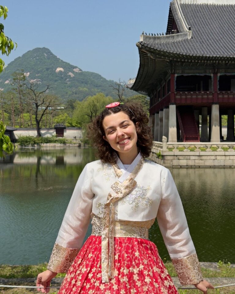A smiling woman wearing a traditional Korean hanbok poses in front of a pond and historic palace building, with green trees and a mountain in the background on a sunny day.