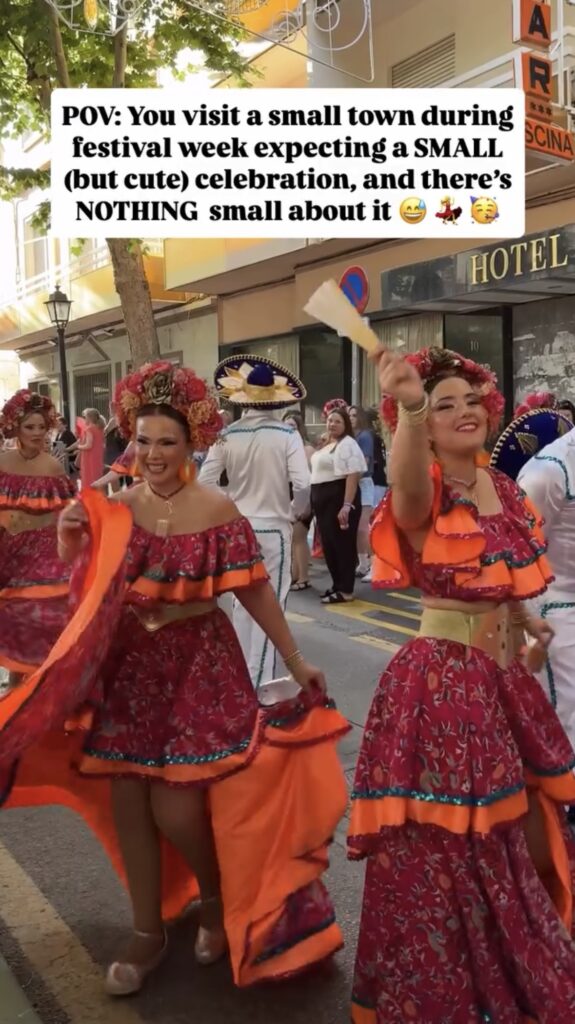 Women in vibrant pink and orange traditional dresses dance joyfully in a lively street parade during a festival, with onlookers and other participants in colorful costumes nearby. A text overlay describes the unexpectedly grand celebration.