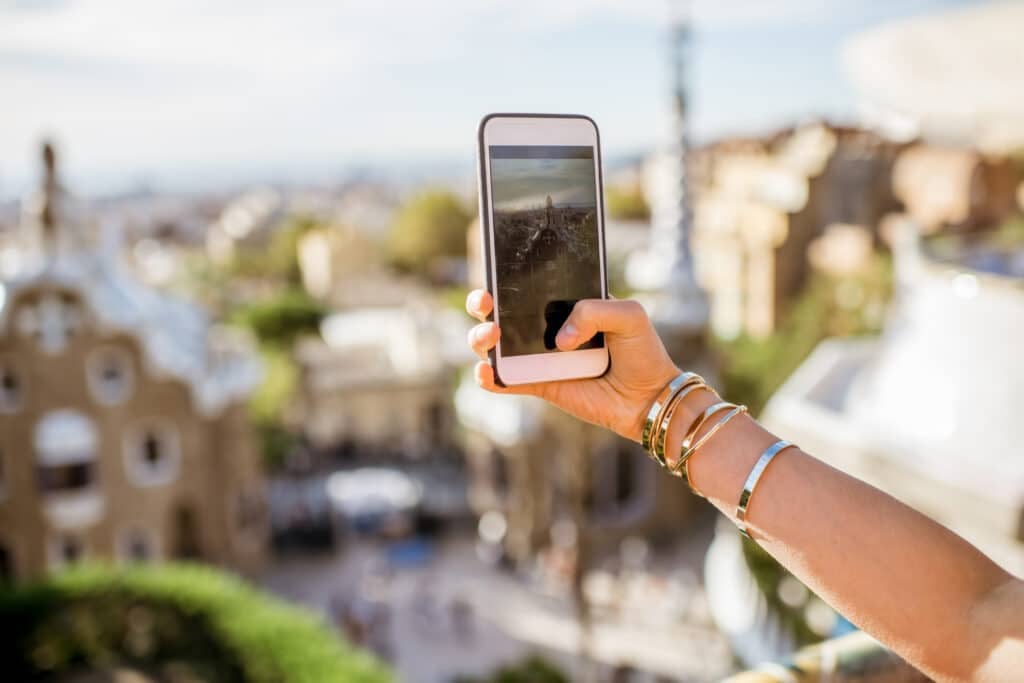A person wearing several gold bangles takes a photo with a smartphone, capturing a scenic, sunlit city view from an elevated point with ornate buildings in the background.