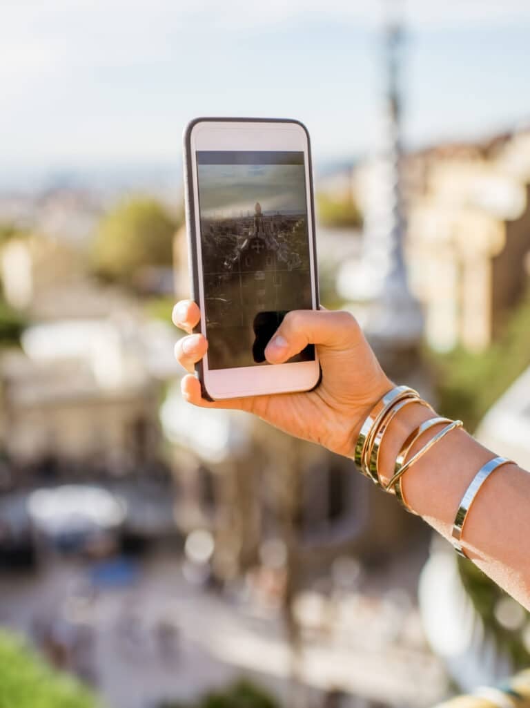 A person wearing several gold bangles takes a photo with a smartphone, capturing a scenic, sunlit city view from an elevated point with ornate buildings in the background.