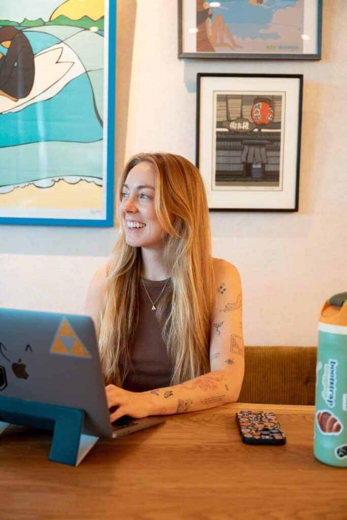 A smiling woman, a travel blogger and creator of Bootstrap Blogging, with long blonde hair works on a laptop at a wooden table. She sits in front of colorful wall art and has a phone and water bottle on the table.