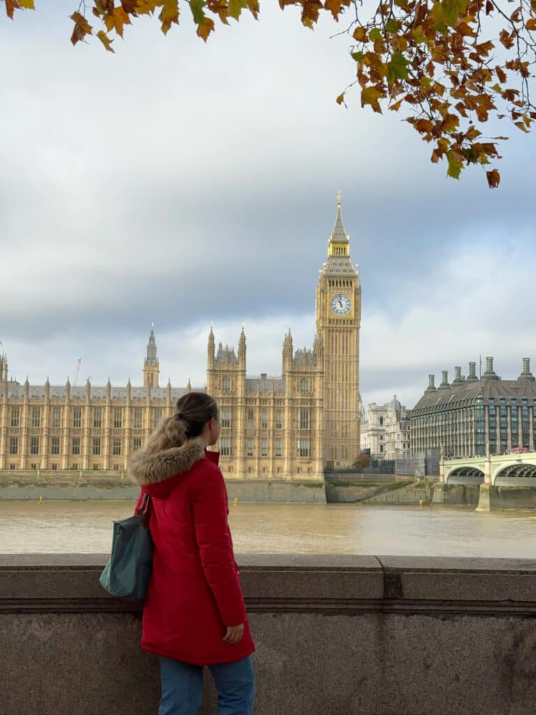 A person in a red coat stands by the River Thames, looking at the Big Ben clock tower and the Palace of Westminster in London, with autumn leaves overhead.
