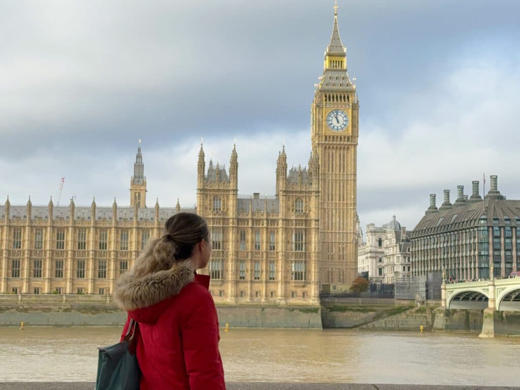 A person in a red coat stands by the River Thames, looking at the Big Ben clock tower and the Palace of Westminster in London, with autumn leaves overhead.