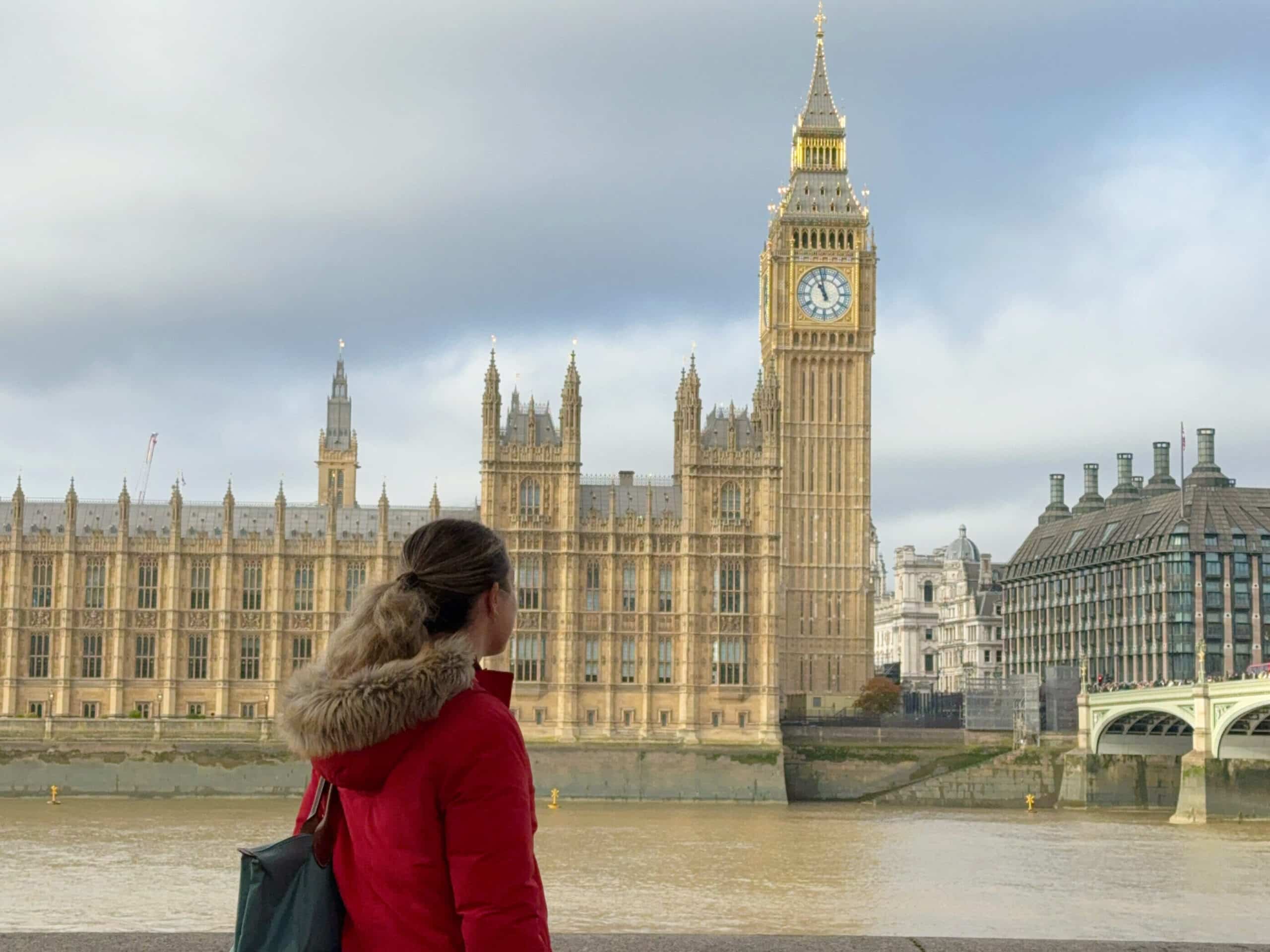 A travel blogger woman in a red coat stands by the River Thames, looking at the Big Ben clock tower and the Palace of Westminster in London, with autumn leaves overhead.