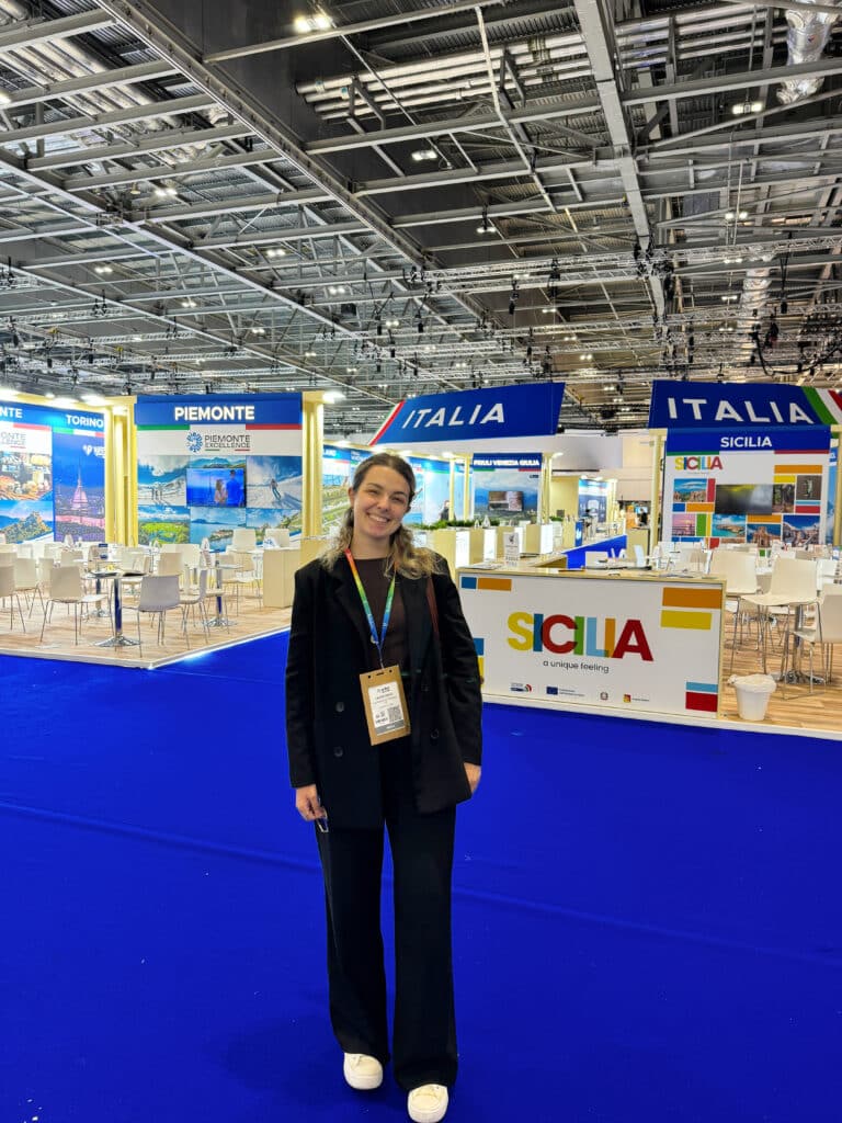 A woman smiles while standing on a blue carpet in front of colorful booths for Sicilia, Italia, Piemonte, and Torino at a large, well-lit exhibition hall. She wears a black outfit and a conference badge.