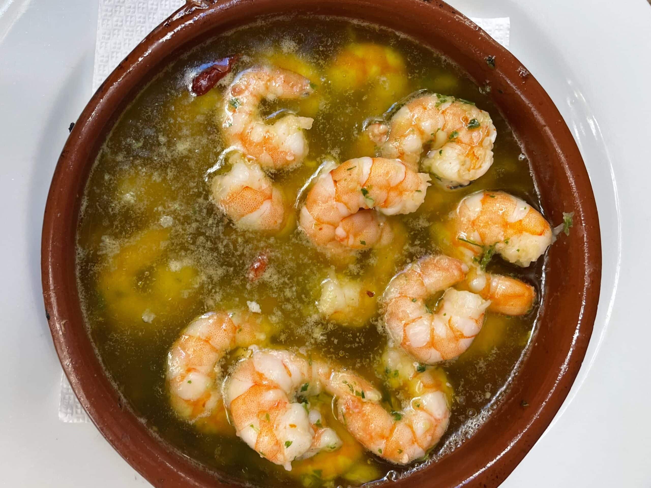 A brown ceramic bowl filled with shrimp cooked in a garlic and hot oil, a classic Madrid food to try, placed on a white plate with a white napkin underneath.