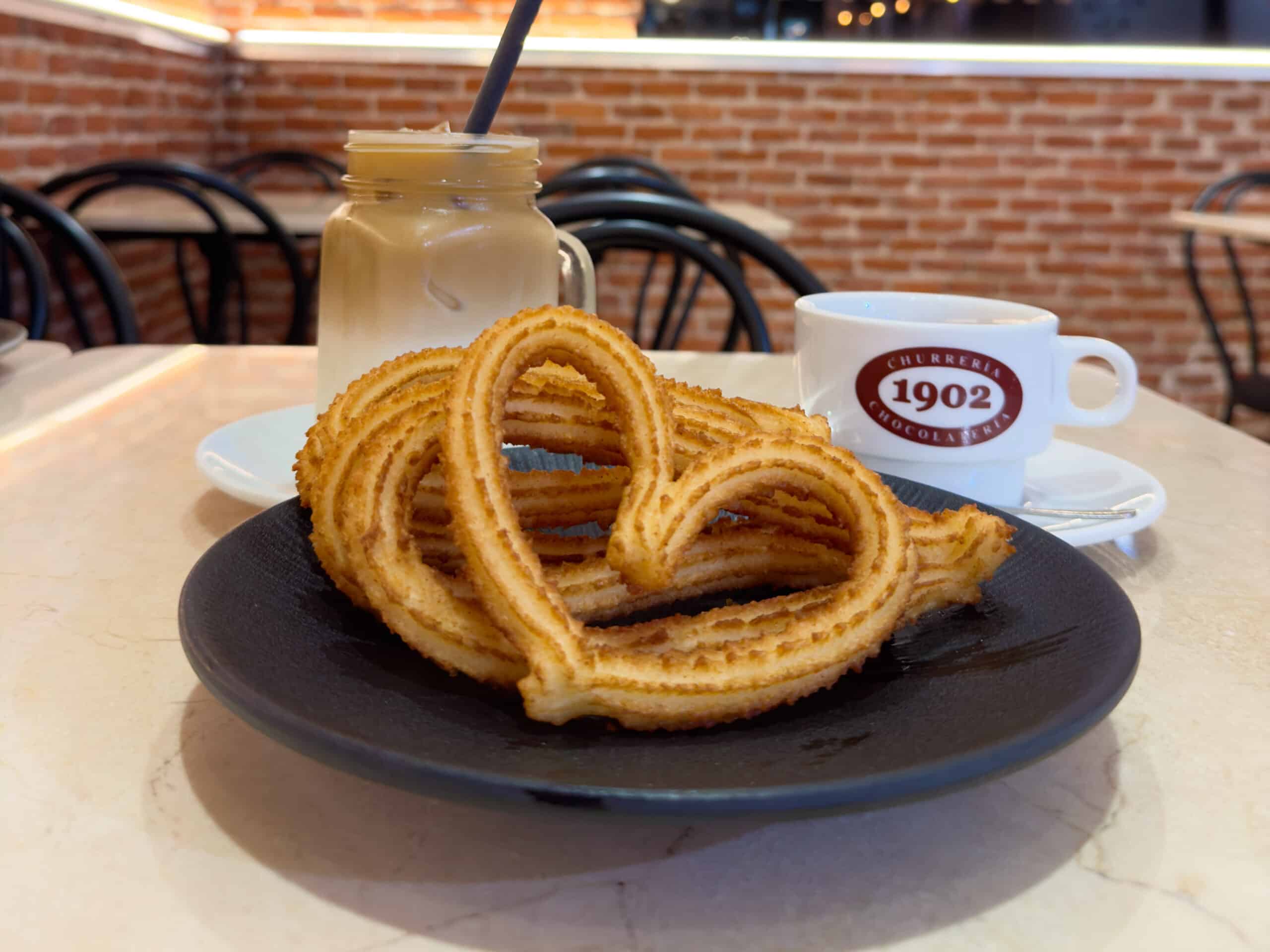 A black plate with two heart-shaped churros is on a table. Behind it, a glass of iced coffee and a cup labeled "Churrería 1902" create the perfect setting for enjoying Madrid traditional food against a cozy brick wall background.