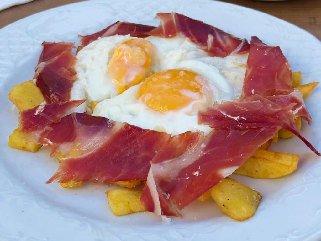 A white plate with French fries topped with two sunny-side-up eggs and thin slices of cured ham, "huevos rotos"—a classic Madrid food to try featured in any authentic Madrid food guide.