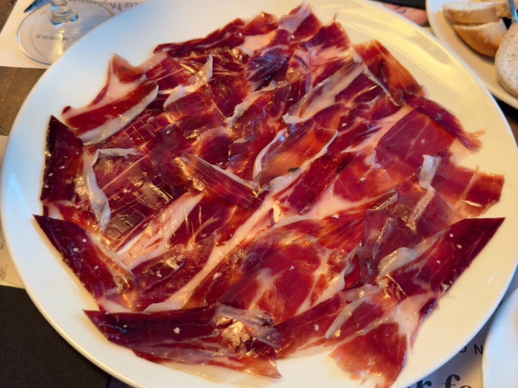 A white plate filled with thin slices of cured Spanish jamón ibérico, showcasing marbled red meat and white fat—a must-have Madrid traditional food. In the background, part of a bread roll and a wine glass are visible.