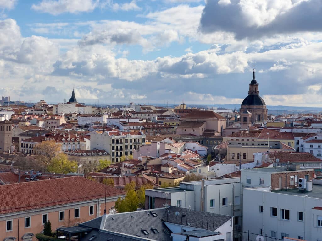 A panoramic view of a European city with red-tiled rooftops, historic buildings, and church domes under a partly cloudy sky.