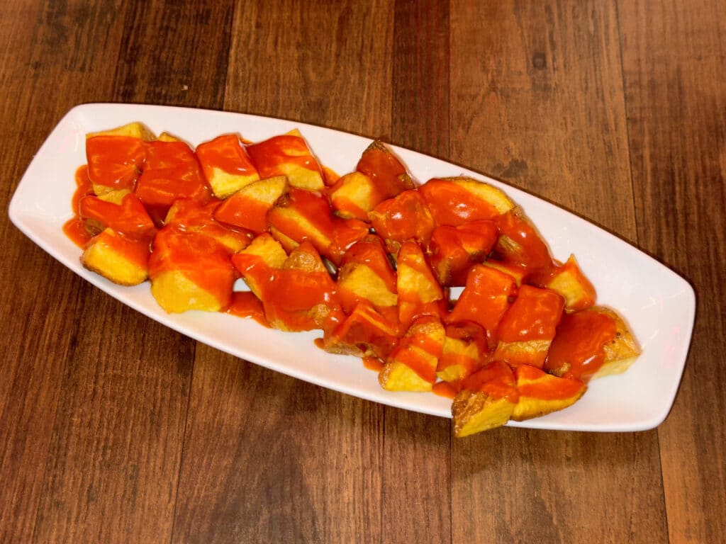 A white oval plate filled Patatas Bravas, crispy potato cubes topped with a generous amount of bright orange-red sauce, a classic example of Madrid traditional food, placed on a wooden table.