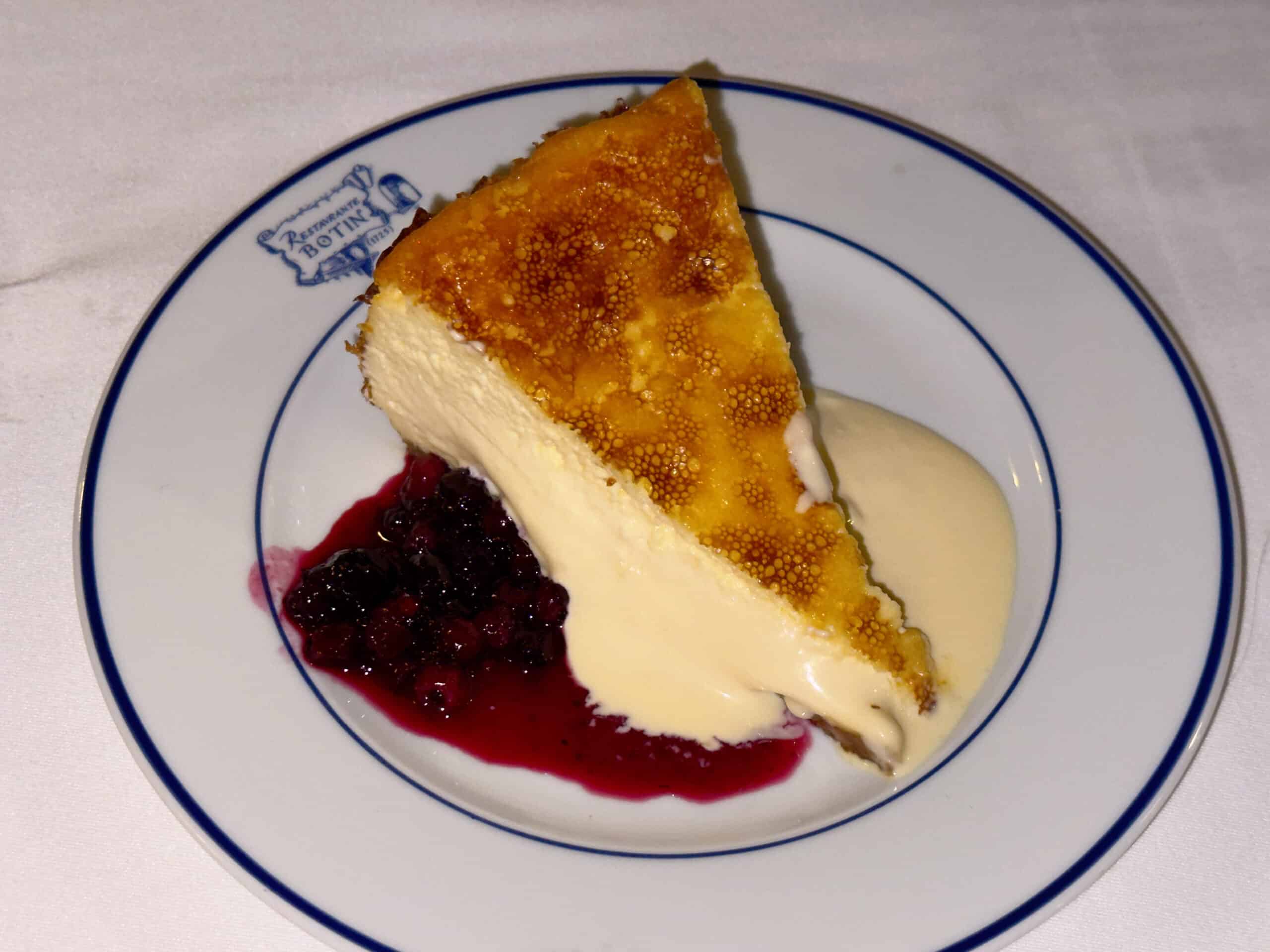 A slice of cheesecake topped with a caramelized layer, served with vanilla sauce and a side of mixed berry compote on a white plate with a blue rim—a must-try dessert featured in any Madrid food guide.