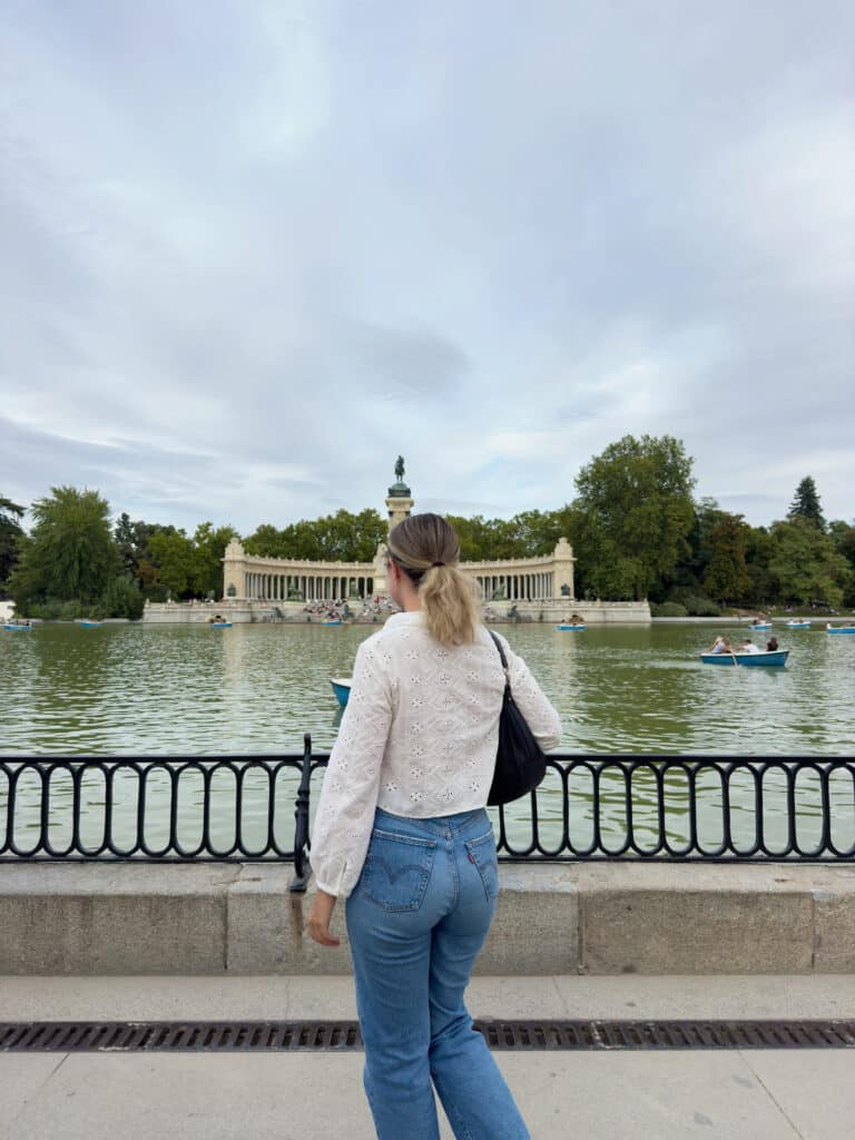 A person with blonde hair in a ponytail, white embroidered top, and blue jeans stands by a lake, facing a monument across the water in a park. Some boats are visible on the lake, under a cloudy sky.