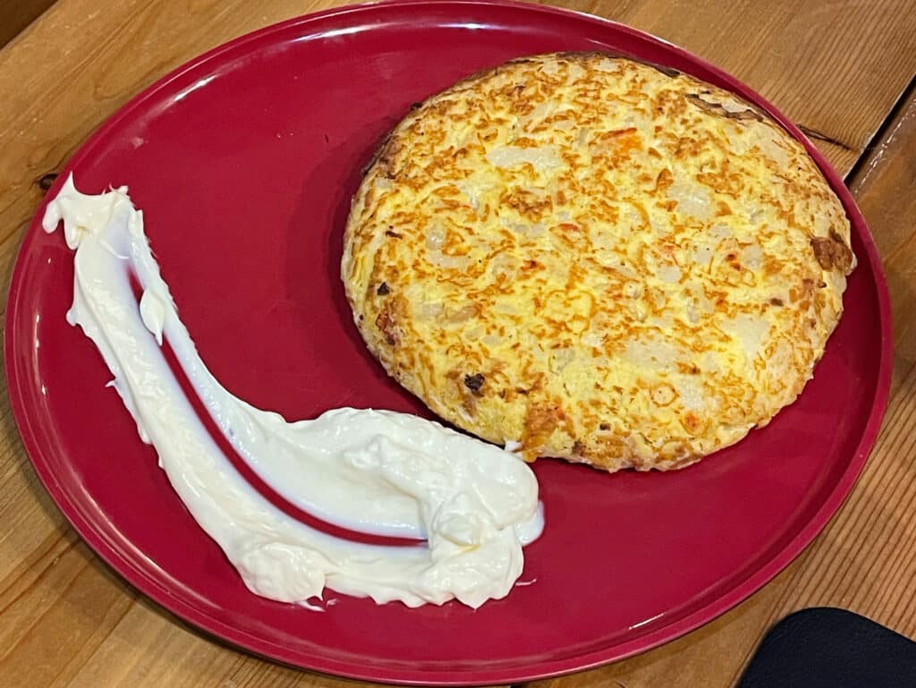 A round Spanish omelette, tortilla española, sits on a red plate next to a swirl of thick white sauce, aioli, on a wooden table—an inviting example of Madrid traditional food.