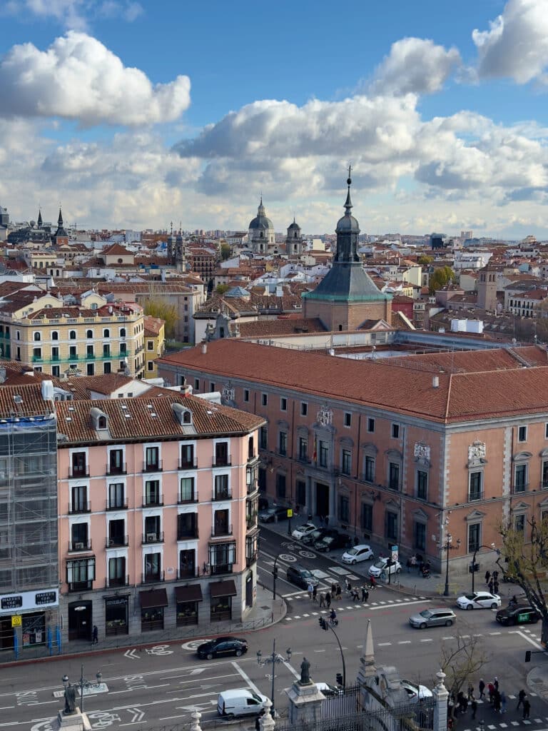 A cityscape view of Madrid featuring historic buildings with red-tiled roofs, church domes, and spires under a blue sky with clouds. Vehicles and people are visible on the streets below.