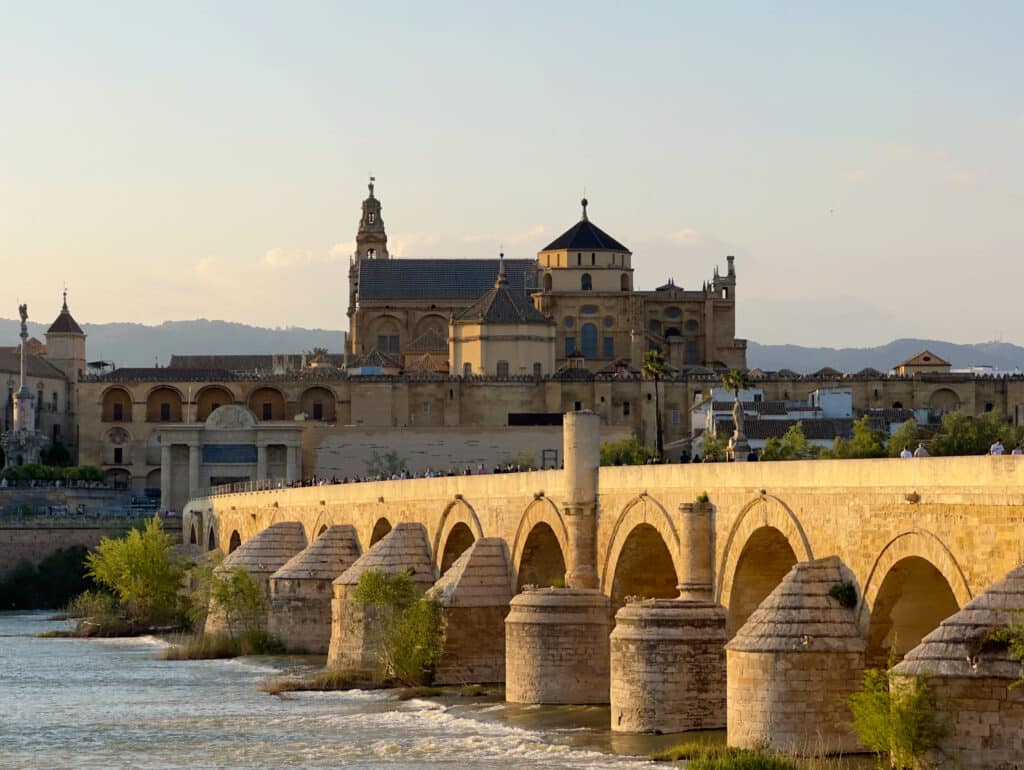 Stone bridge with a row of arches crossing a river, and a historic city with domed towers in the background under a pale blue sky.