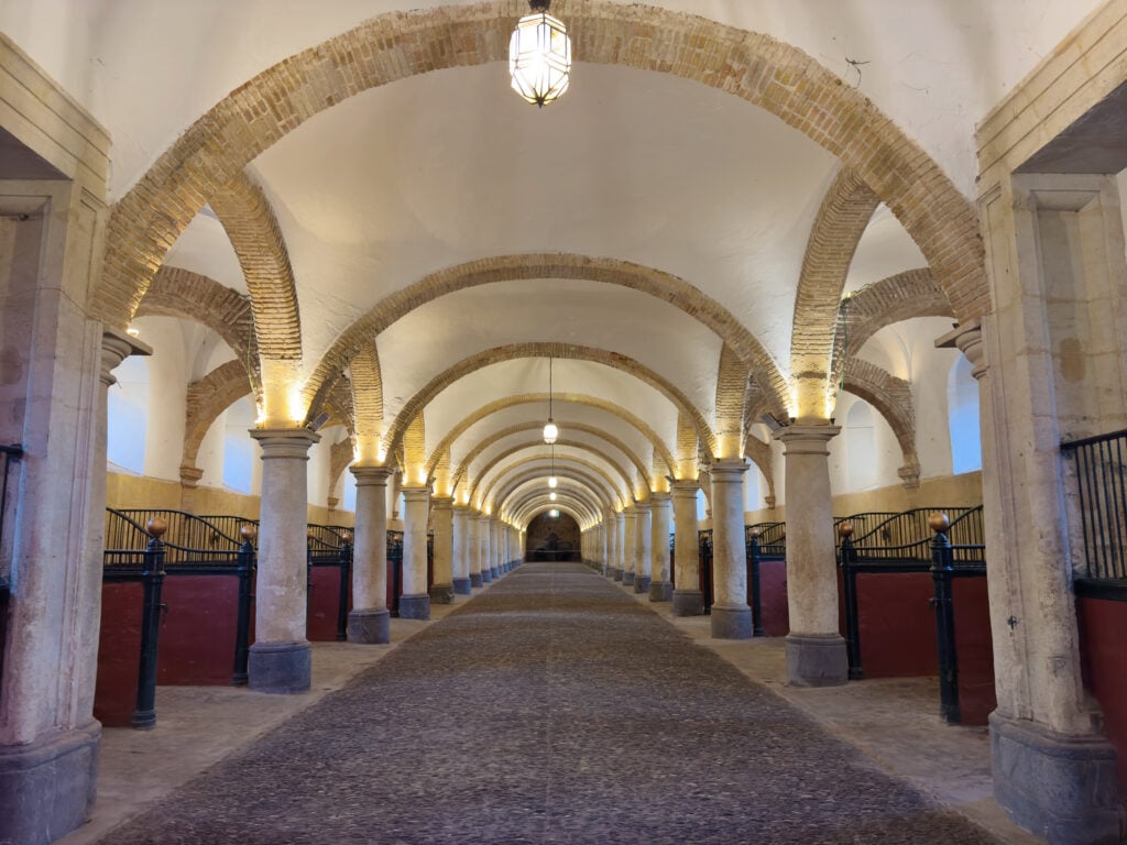 A long, arched hallway with stone columns and illuminated arches, featuring symmetrical horse stalls on both sides and a cobblestone floor, leads into the distance under hanging lanterns. Royal Stalls of Cordoba