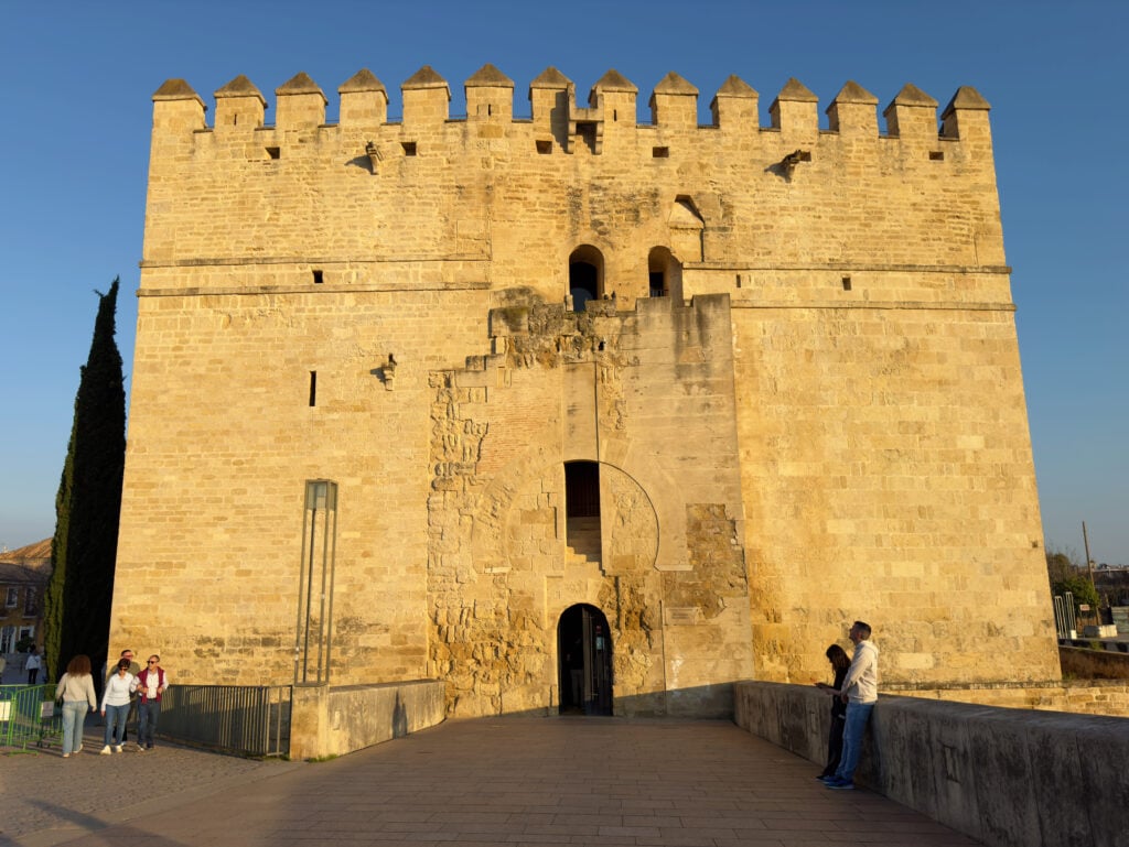 A large, historic stone fortress with crenellated battlements, illuminated by sunlight. A few people stand and walk near the entrance at the base of the structure, and a tall cypress tree is visible to the left.
