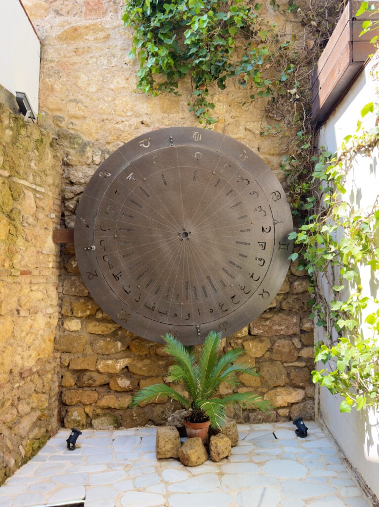 A large round sundial with hour markings and symbols mounted on a stone wall, surrounded by greenery, with a small potted palm plant at its base and white tiled floor below.