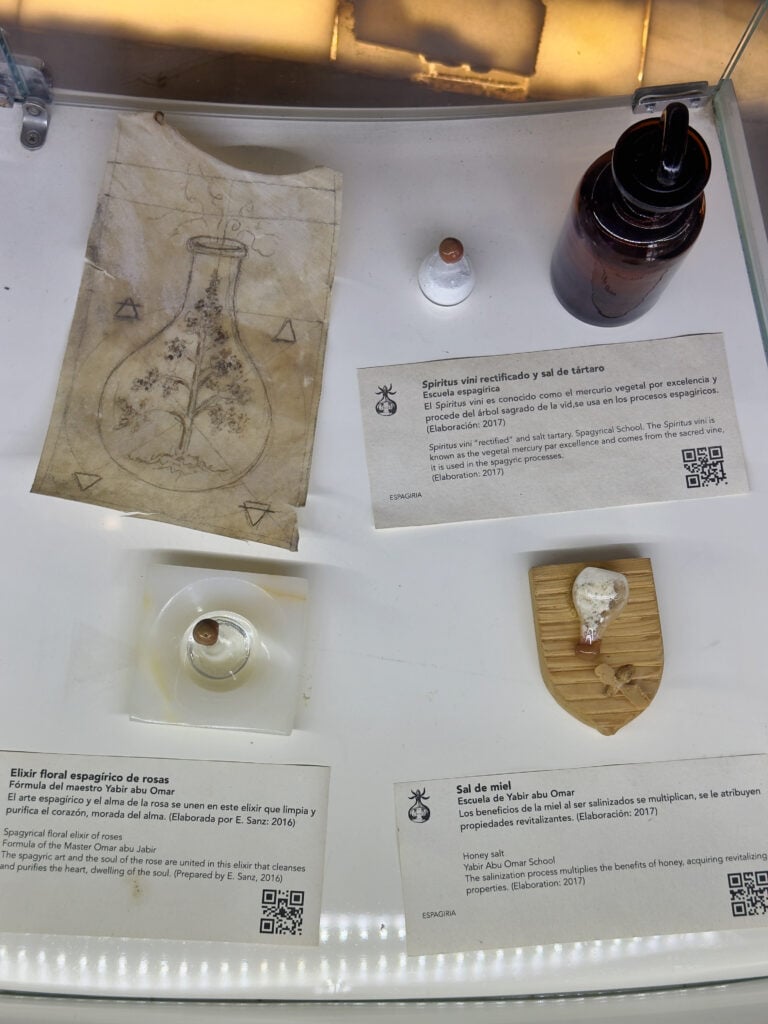 Display shelf with a parchment sketch of a plant in a flask, a small glass vial, and a tall brown bottle beside an informational card in Spanish about esparigiria/elixirs in the Casa de Alquimia in Cordoba