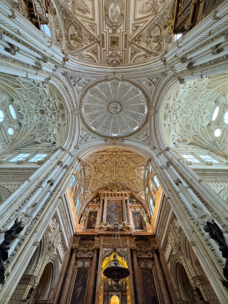 Ornate cathedral interior viewed upward, showing an intricate, vaulted ceiling with detailed carvings and frescoes above an elaborate altar adorned with religious artwork and statues. Light streams in through high windows.